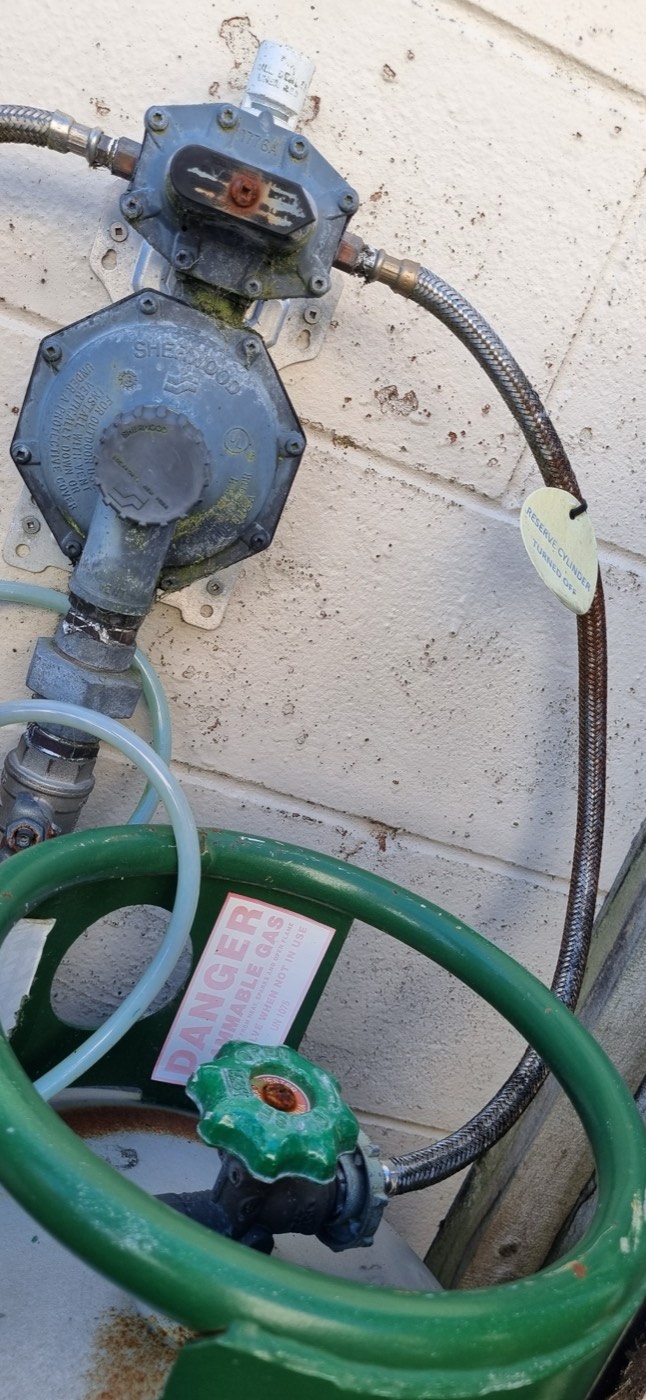 An old, weathered and corroded LPG changeover regulator on an outside wall above two gas bottles